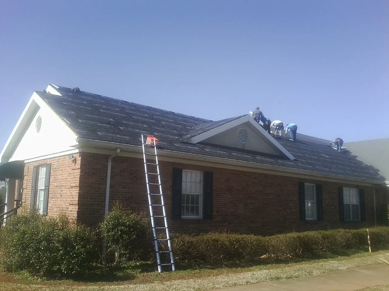 Crew tearing shingles off a brick ranch roof with a ladder set — Metal Roof Repair work by our crew in Slater